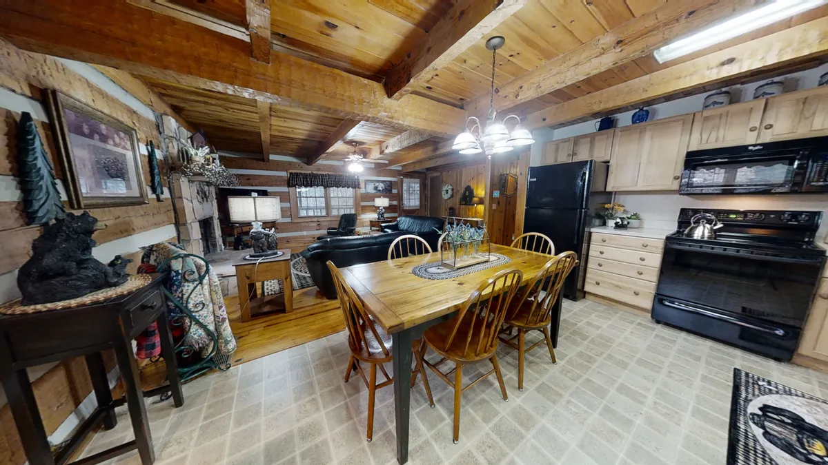 An open-plan view highlighting the dining table set for a meal, visible kitchen appliances, rustic wooden beams, and part of the living area. The arrangement shows seamless flow between the kitchen and living space, emphasizing openness and togetherness. The cozy ambiance hints at family meals or friendly gatherings.