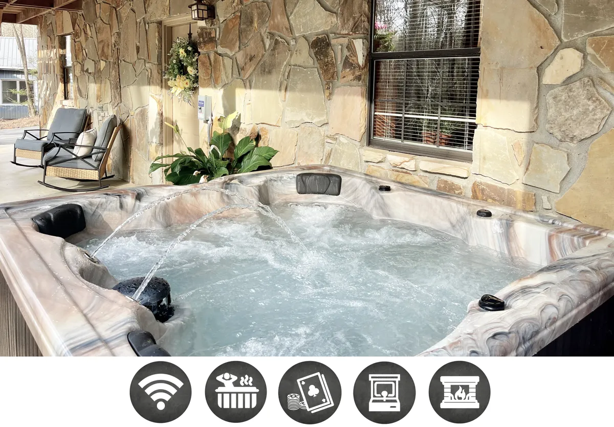 The image features a cozy indoor seating area adjacent to a bubbling hot tub, set against a rustic stone wall. Large windows frame views of the surrounding Smoky Mountain woods, and plush rocking chairs invite guests to relax and soak in the peaceful, semi-private atmosphere.