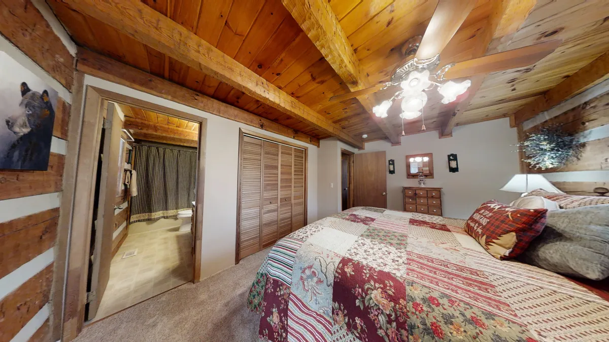A charming bedroom featuring a patchwork quilt on the bed, rustic wooden beams overhead, bear-themed artwork, and a dresser with decorative items. The window invites natural light, enhancing the connection to the forest surroundings.