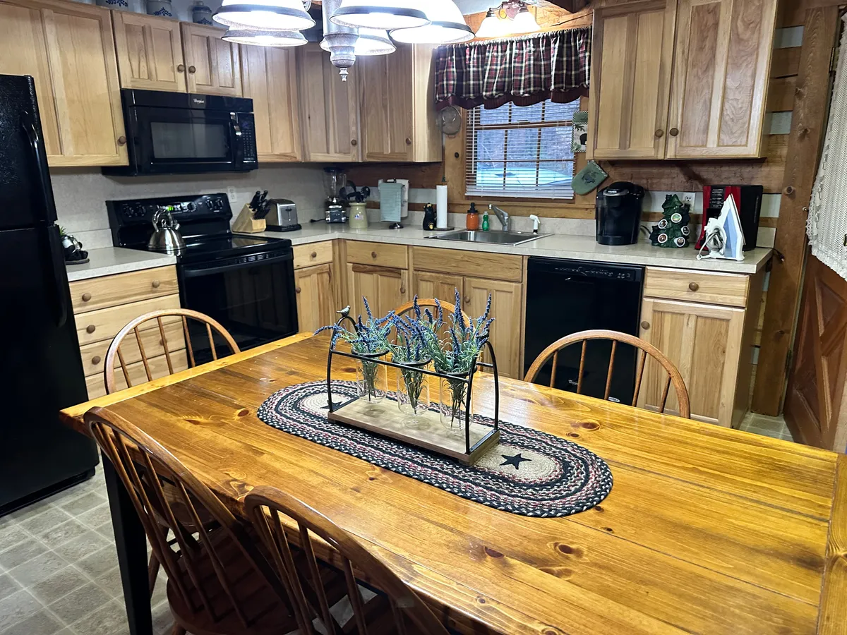 A view of the kitchen and dining area featuring a sturdy wooden dining table, sleek black kitchen appliances, ample cabinetry, and a decorative centerpiece. The warm wood tones and inviting lighting create a homely, social atmosphere. The kitchen setup appears ready for group meals, cooking, and entertaining guests, with visible counter space and modern amenities.