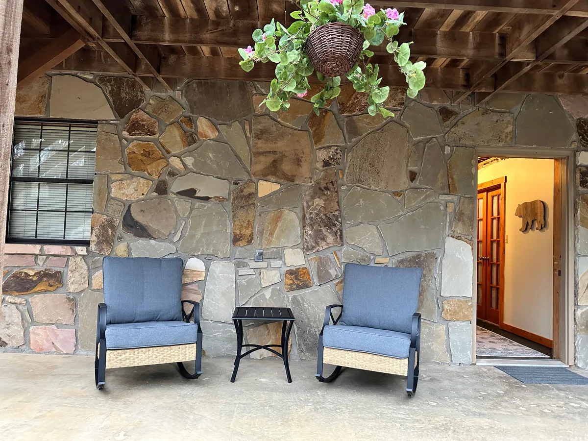 A cozy porch area with two comfortable chairs set around a small black table, complemented by a hanging plant. The porch is surrounded by lush greenery, with sunlight filtering through the leaves, creating a tranquil and private outdoor retreat.