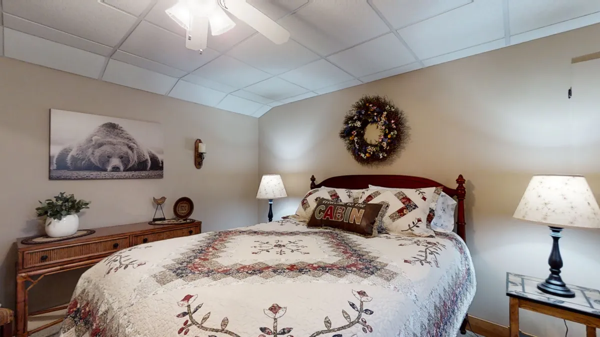 This interior image showcases a welcoming bedroom featuring a bed with a cozy quilt, bear-themed artwork that connects to the cabin's mountain setting, a decorative wreath above the bed, and nightstands with warm lighting from lamps. The overall mood is tranquil and inviting, emphasizing comfort and nature-inspired decor.