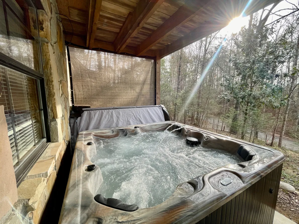 An outdoor hot tub nestled under a wooden structure with natural surroundings, promising a tranquil escape after indoor fun. The setting invites relaxation under the trees, with year-round usability and sunset views.