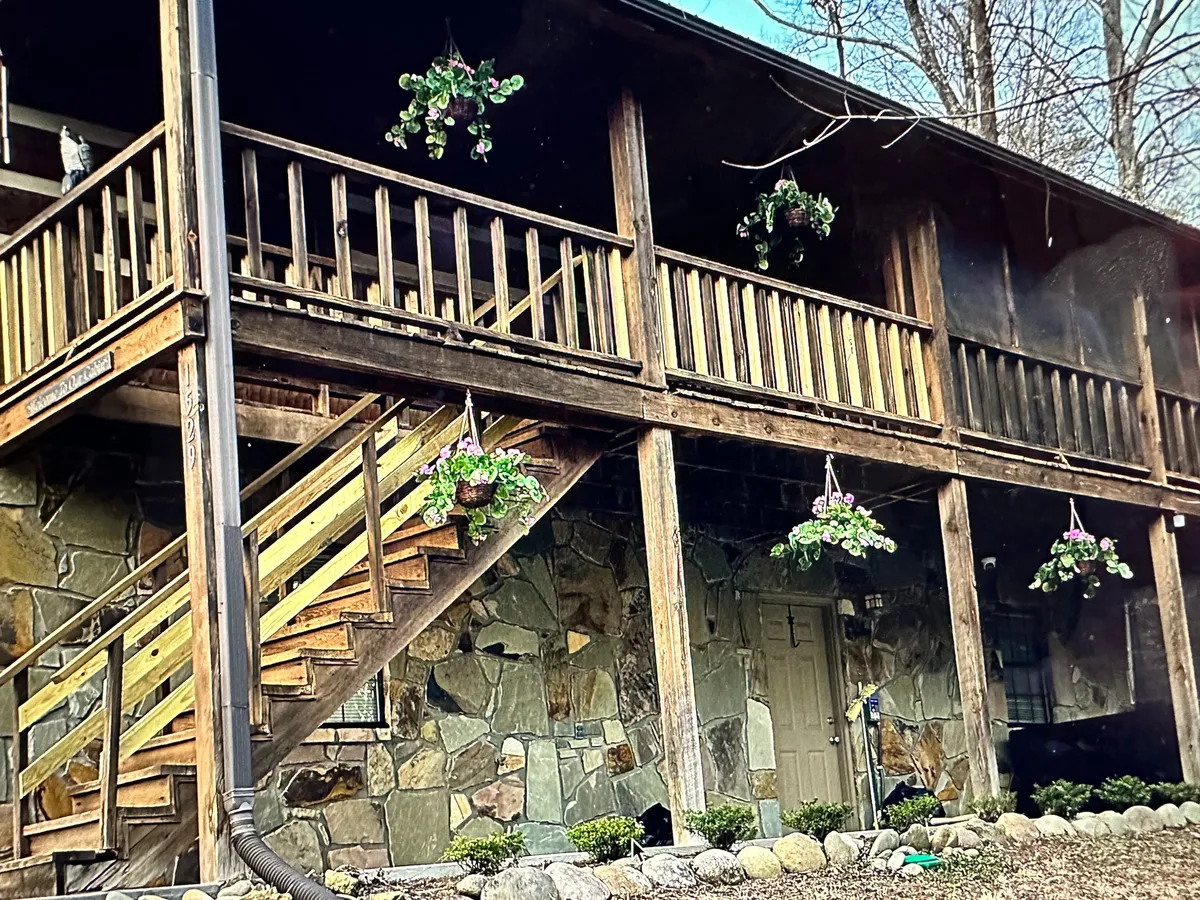 This inviting exterior image captures the entrance to the property, featuring wooden stairs, hanging flower pots, a stone wall, and a welcoming door. The porch is framed by natural foliage, creating a perfect setting for group gatherings and outdoor enjoyment.