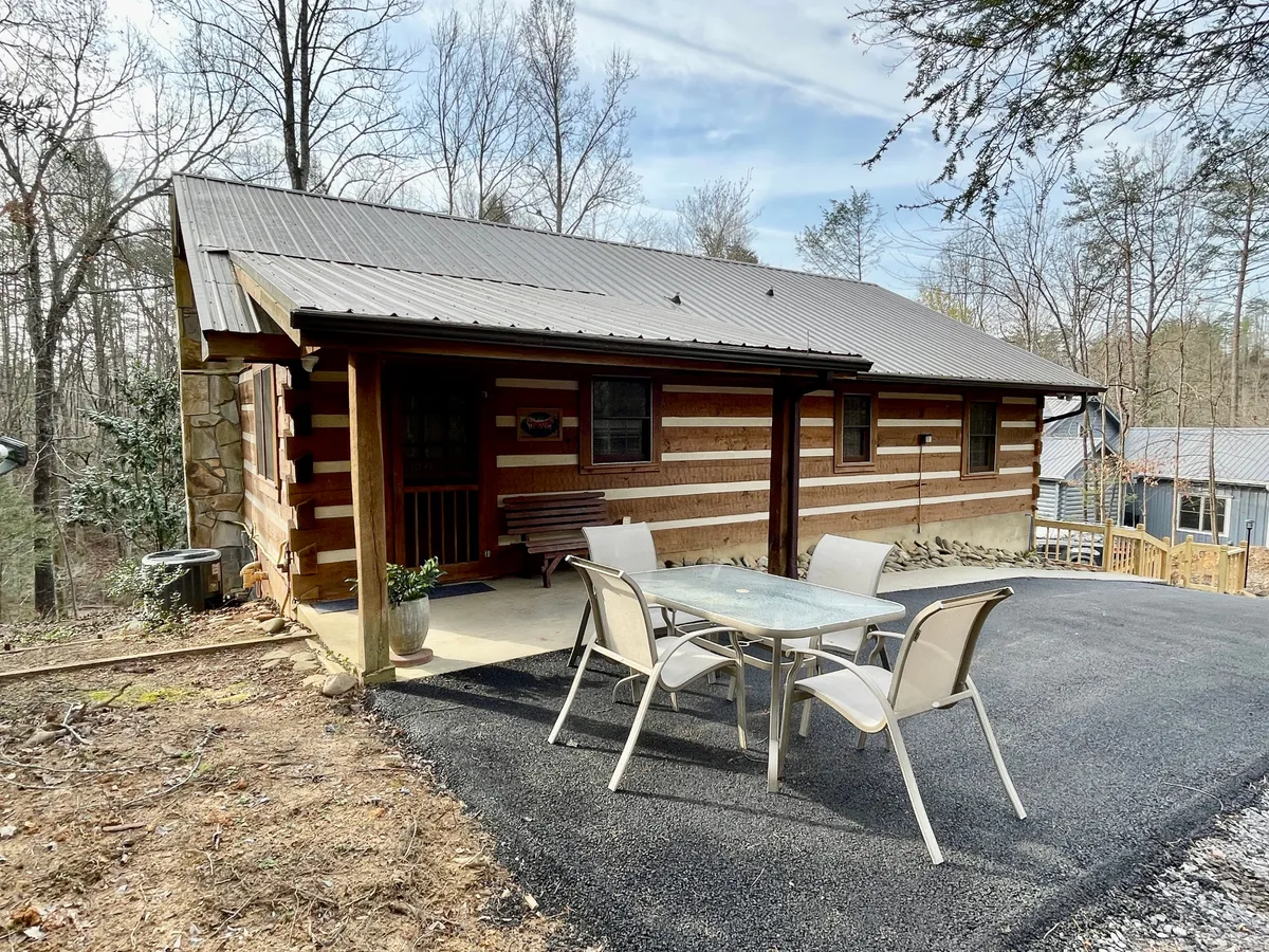 The image shows the inviting exterior of a log cabin, surrounded by lush trees and featuring an outdoor seating area. The natural setting creates a sense of tranquility and connection to the Smoky Mountains, encouraging guests to relax after a day of adventure.
