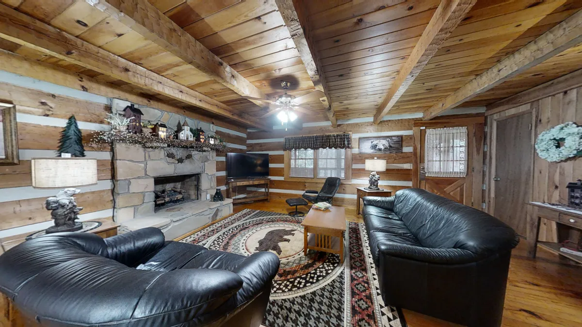 This view focuses on the living room's warm, rustic charm with leather sofas arranged around a grand stone fireplace, wooden beams overhead, a decorative rug, and a TV. The mood is inviting and perfect for relaxing, reading, or enjoying time together.