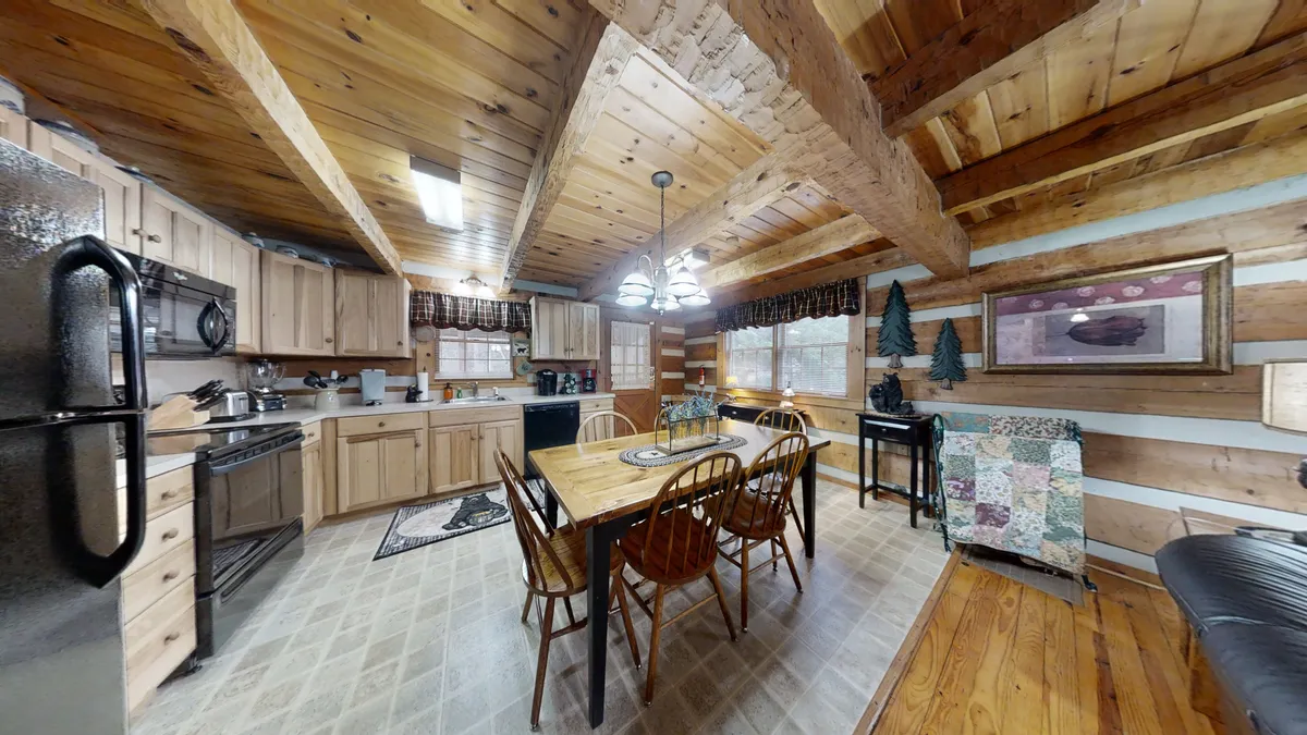 This interior view highlights a wooden dining table set for six, adjacent to the kitchen area, all beneath exposed wooden beams and with large windows offering woodland views. Natural light fills the space, emphasizing both the dining and kitchen areas as social hubs.