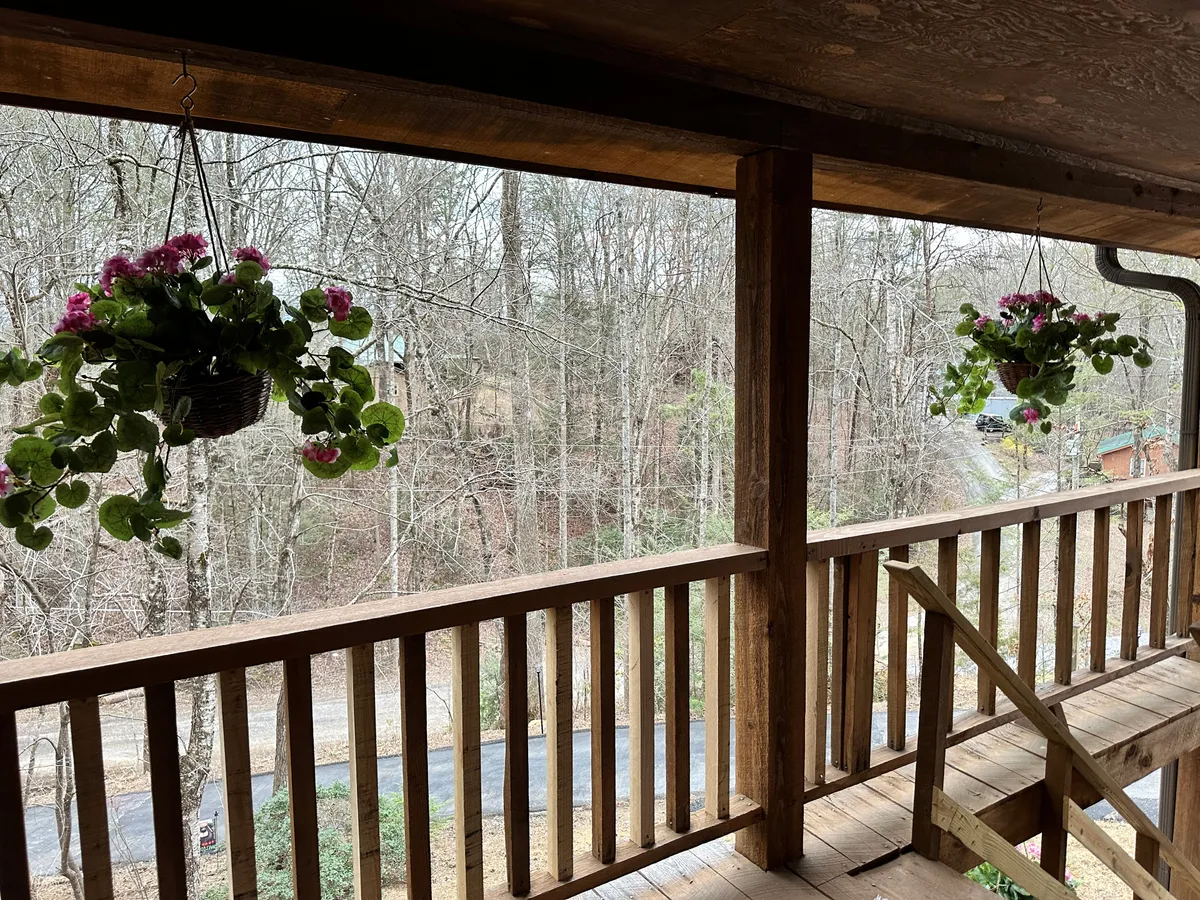 This exterior image showcases hanging flower baskets, a classic wooden railing, and a lush, natural landscape. The scene is tranquil, with soft sunlight filtering through surrounding trees, evoking seasonal changes and inviting outdoor relaxation.