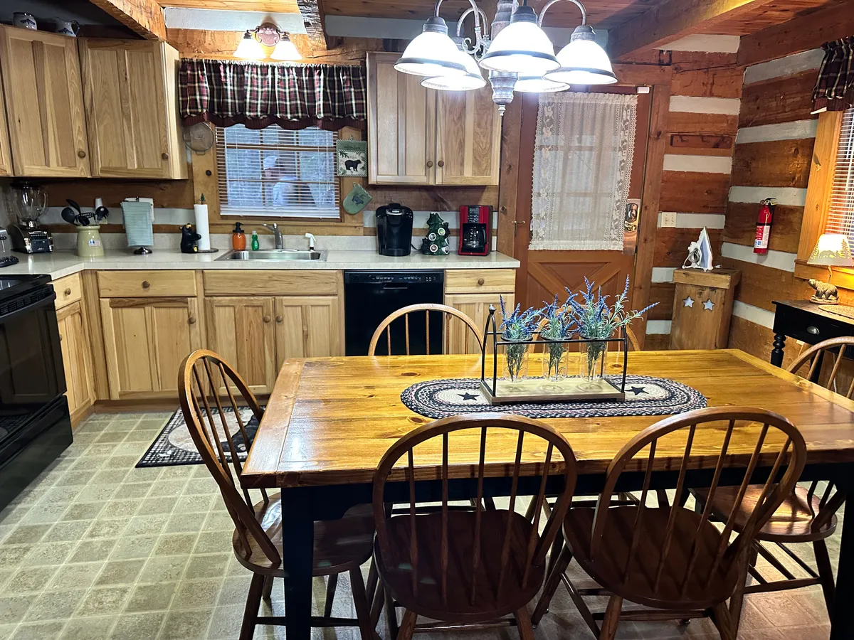 A warmly lit kitchen showcasing wooden cabinets, kitchen appliances, a window with plaid curtains, and a wooden dining table. The scene feels intimate and functional, ready for cooking or gathering. The traditional styling and inviting light capture the homely, welcoming spirit described in the section.