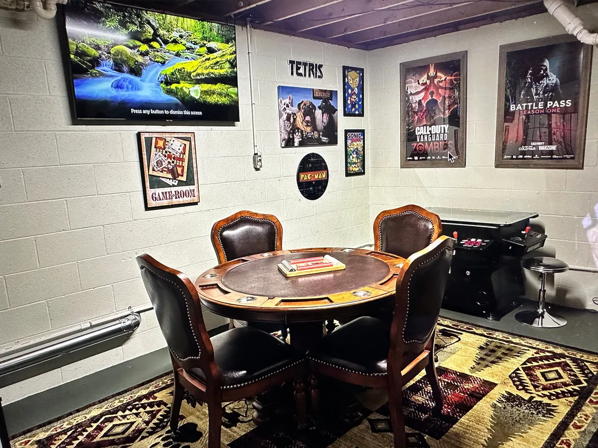 A playful downstairs game room featuring a round wooden table surrounded by leather chairs, a television screen perfect for movie viewing, game-themed posters on the walls, and a bar-style gaming table. The space exudes a relaxed yet energetic vibe, ideal for group activities, board games, and game competitions.