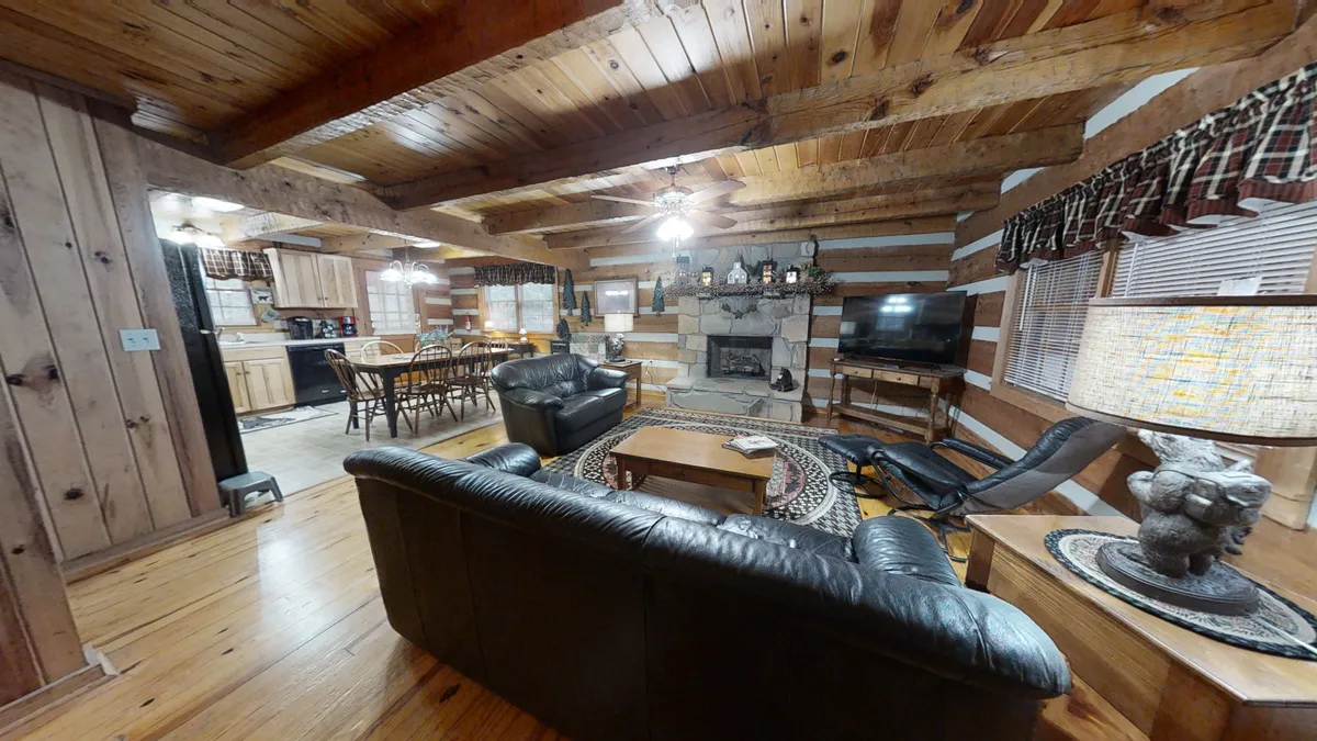 This image features the main living room with a leather sofa set, a stone fireplace, exposed wooden beams, a TV, and a glimpse of the adjacent dining area. The lighting is warm and natural, and the space feels both spacious and inviting, perfect for gatherings and relaxation.